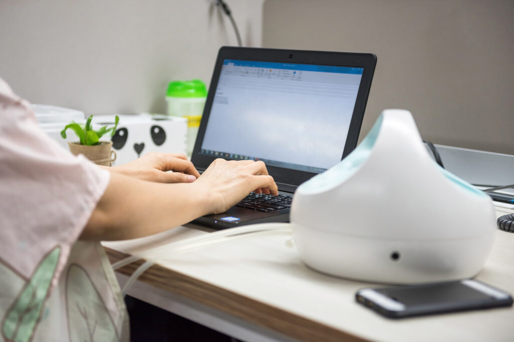 Busy mother pumping breastmilk by Automatic breast pump machine wirh Nursing or Breastfeeding fabric cover while working on laptop pc computer on table. Motherhood in corporate office.