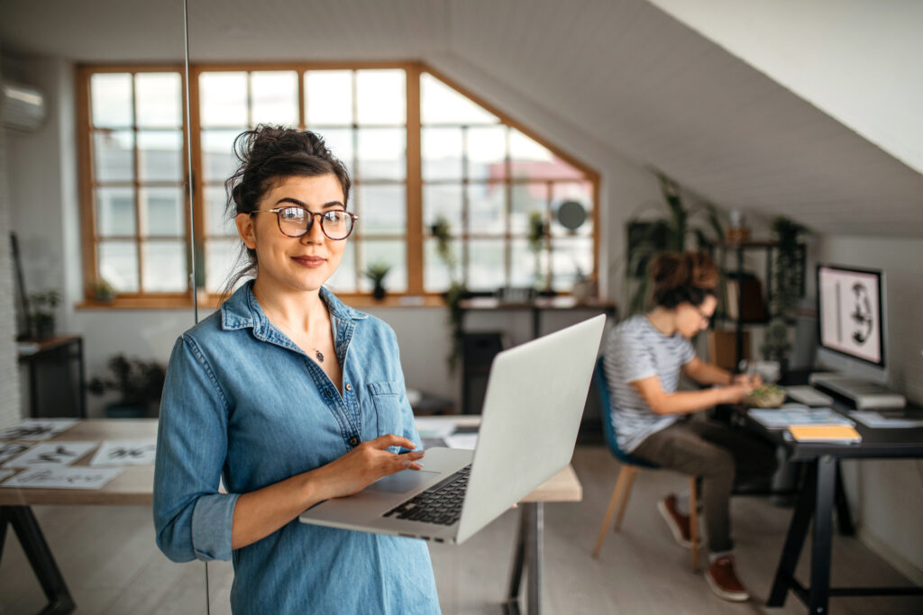 Portrait of young smiling woman in creative office, standing, holding laptop, looking at camera.