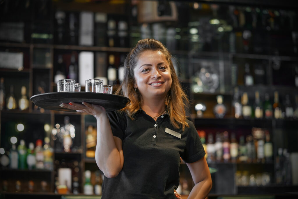 Waitress carries a whiskey glass on a tray in hotel restaurant, bar. The concept of service. Shelves with bottles of alcohol in the background. Night time.