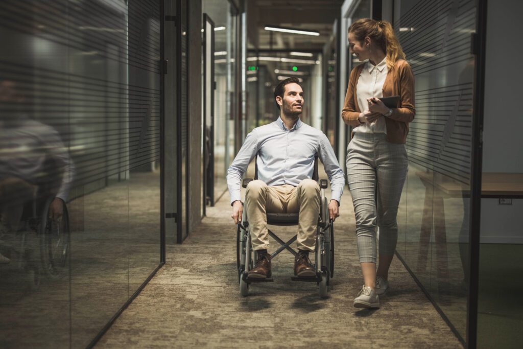 Happy businessman in a wheelchair and his female colleague communicating while being on the move in a hallway.